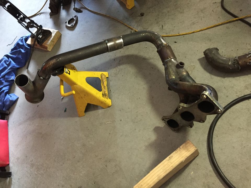 Custom exhaust manifold and downpipe assembly on concrete floor with yellow jack stand.