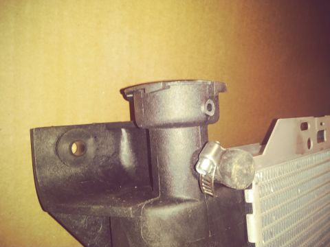 1996 Ford Bronco - A close-up view of a black plastic radiator neck with a metal hose clamp attached to a rubber hose. The radiator fins are visible to the ri…