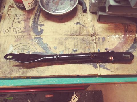 1996 Ford Bronco - A black painted metal bracket is laid out on a workbench with various markings and paint splatters. The bracket has two eyelet holes at eac…