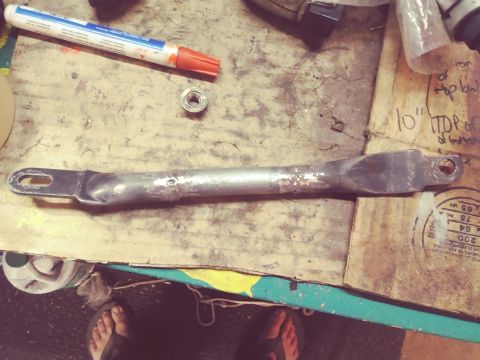 1996 Ford Bronco - A metal bracket with rust and some dents is laid out on a workbench next to a marker and a nut. The bracket appears to be a part of a vehic…