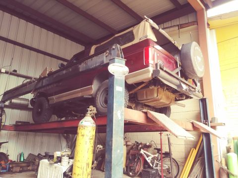 1996 Ford Bronco - A maroon Ford Bronco is raised on a car lift inside a workshop. The vehicle has a tan soft top and a spare tire mounted on the rear.