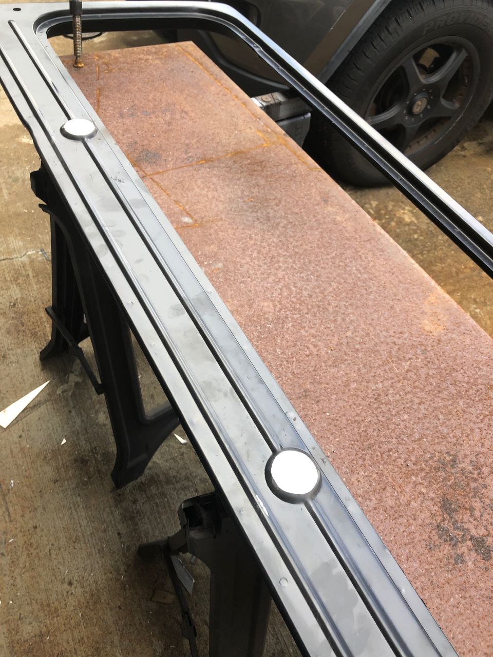 Black trim piece with white inserts on sawhorses, rusty metal, and vehicle tire.