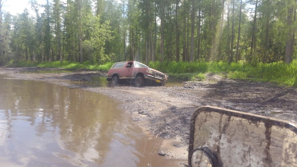 Red SUV stuck in mud and water on a dirt road with trees in the background.