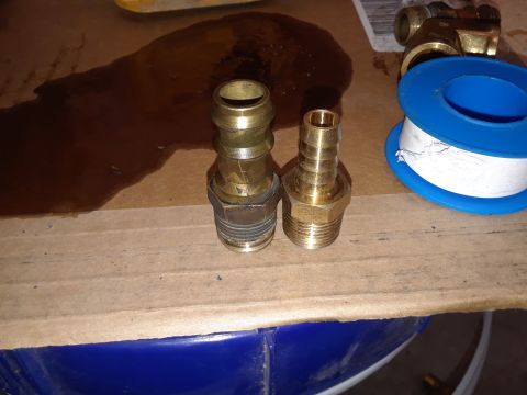 1985 Ford Bronco - Two brass hose barb fittings and a roll of Teflon tape are laid out on a surface with a spill of dark liquid.