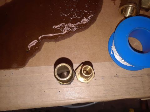 1985 Ford Bronco - Two brass fittings and a roll of blue Teflon tape are on a cardboard surface with a dark liquid spill.