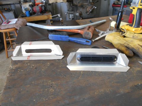 1992 Ford Bronco - Two custom aluminum brackets are on a workbench. One bracket has a rectangular opening, and the other has a black vent with horizontal slat…
