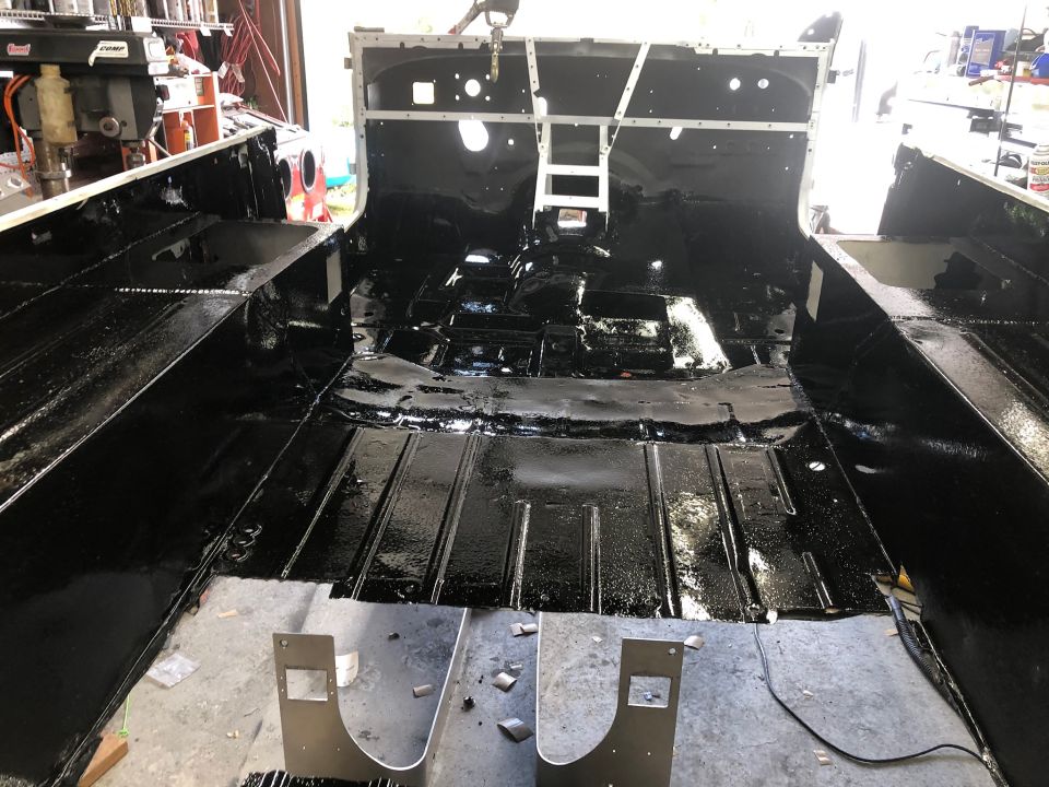 Interior of a Jeep CJ with black undercoating applied to the floor pans and body tub. Some metal brackets are visible in the foreground