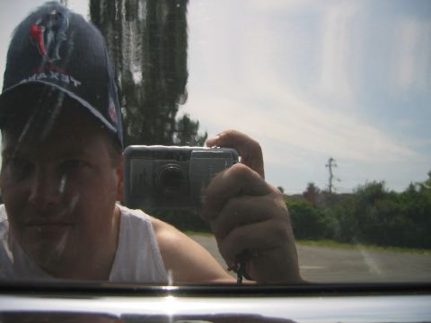 2002 Ford Taurus - Man wearing a dark cap and white tank top photographing his reflection with a silver compact camera in vehicle glass outdoors.