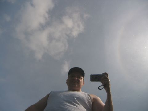 2002 Ford Taurus - Low-angle selfie of a man in a cap and white tank top holding a camera, with a sun halo in the hazy sky behind him.