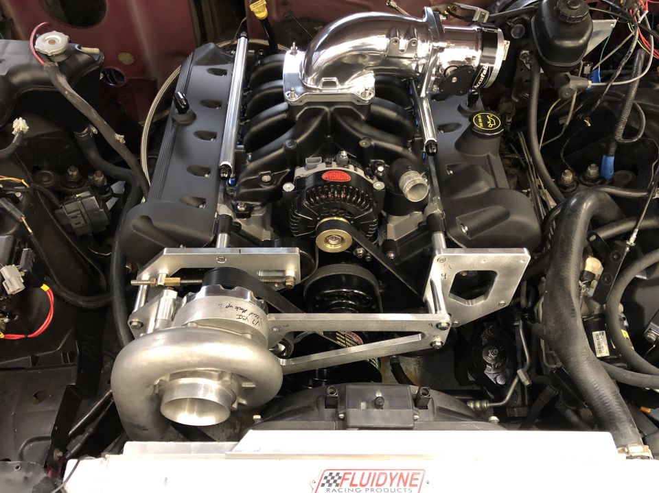 Supercharged V8 engine with black intake manifold and chrome piping. Fluidyne Racing Products logo visible