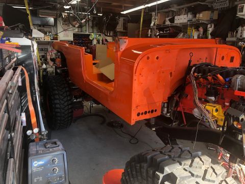 1992 Jeep Wrangler - The body of an orange Jeep CJ is seen in a workshop, with the engine exposed and large off-road tires attached, indicating a restoration pr…