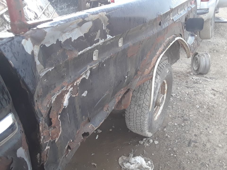 Heavily rusted black truck bed and rear fender with peeling paint and visible holes. A spare tire is on the ground