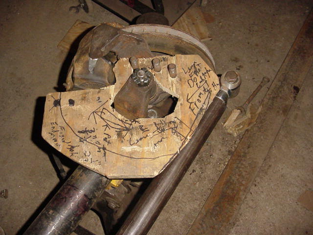 Close-up of a modified axle housing with a wooden jig covered in scribbled notes and measurements. A metal rod extends from the housing