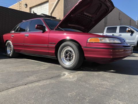 1992 Ford Crown Victoria - A red 1992 Ford Crown Victoria with its hood up and drag wheels is parked outside.