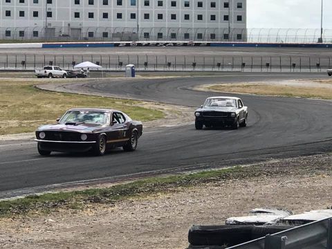 1970 Ford Mustang - Two classic Ford Mustangs compete on a race track with a large building in the background.