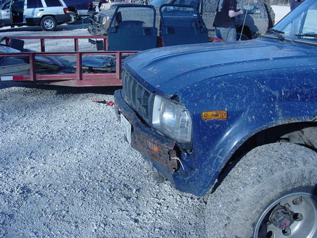 Close-up of a blue pickup truck's front fender, headlight, and tire. A red trailer is visible behind it