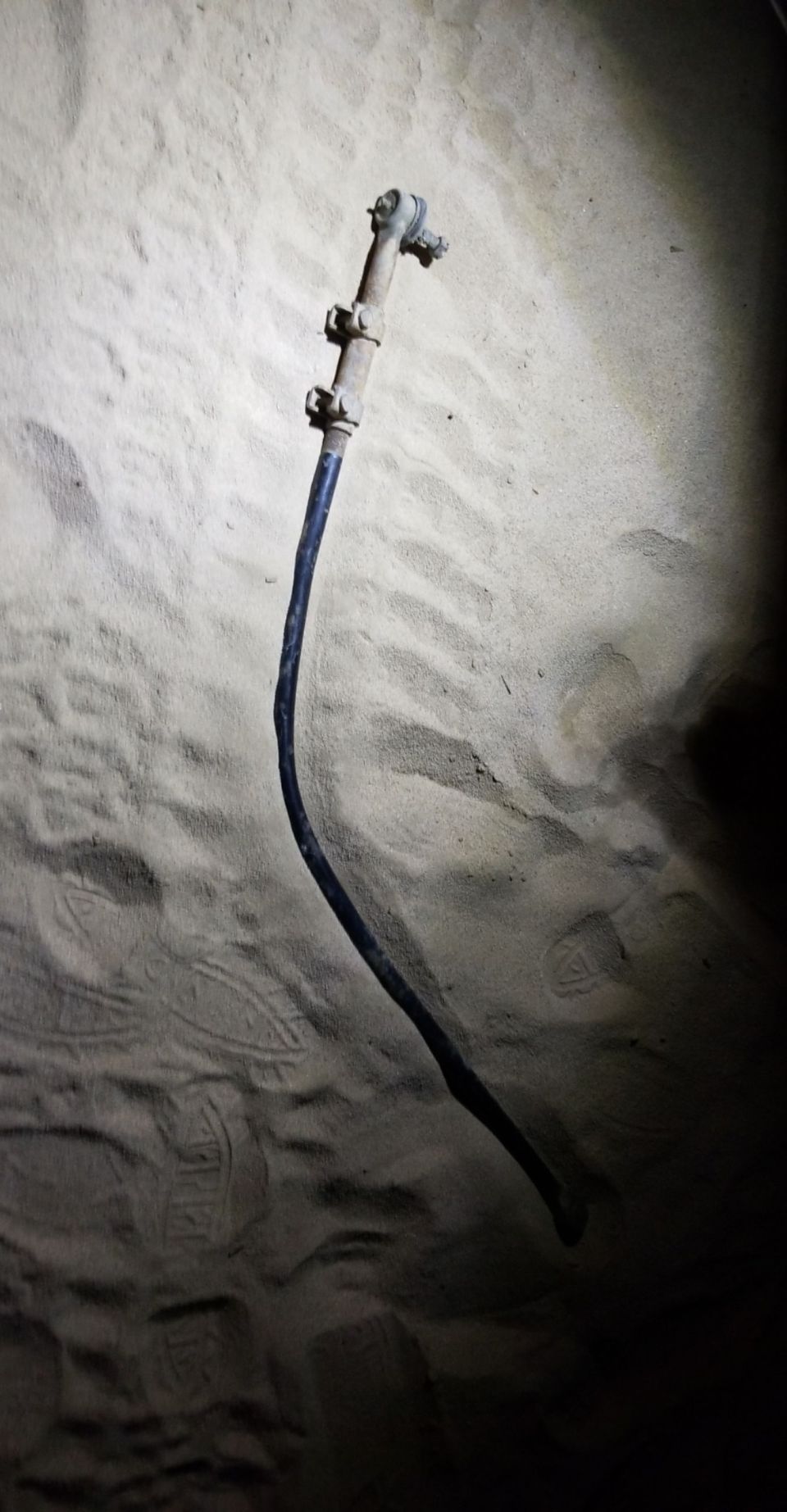 Steering linkage part on sandy ground. The part is metallic with a black hose attached