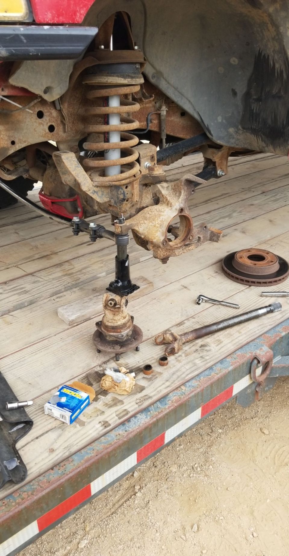 Disassembled front suspension and CV axle on a trailer deck. Red body panel visible