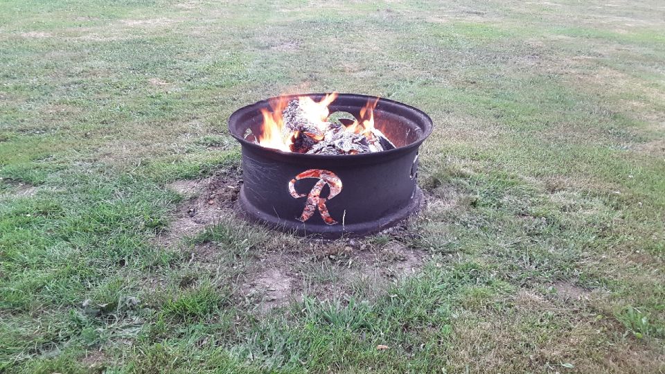 Fire pit made from a tire rim with a cutout 'R' logo, burning wood in a grassy yard