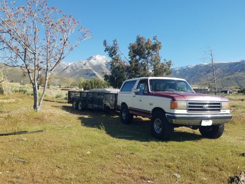 1990 Ford Bronco - A two-tone Ford Bronco is parked in a field, hitched to a trailer, with snow-capped mountains visible in the distance.