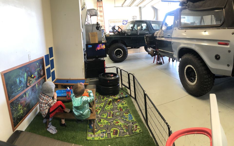 Two Ford Broncos, one gray and one dark green, are parked in a garage. The gray Bronco is on jack stands. Two children play in a fenced-off area