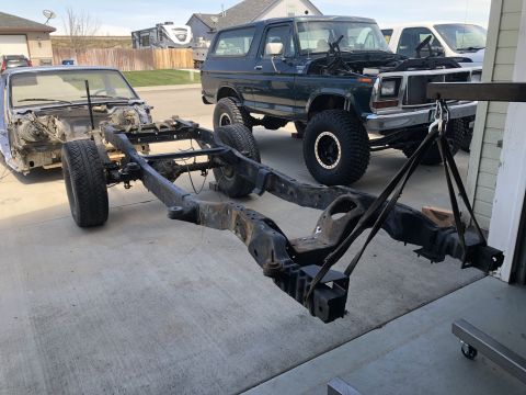 1979 Ford Bronco - A vehicle frame is suspended by a hoist, with a Ford Bronco and a car body in the background.