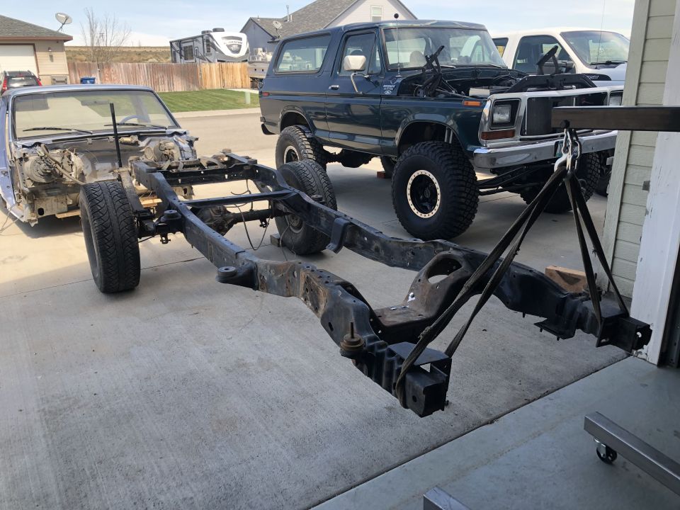 Vehicle frame being lifted by a hoist, with a dark green Ford Bronco and a disassembled car in the background