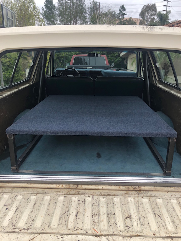 View into the back of a Ford Bronco with a blue carpeted platform bed installed. Another red truck is visible through the windshield