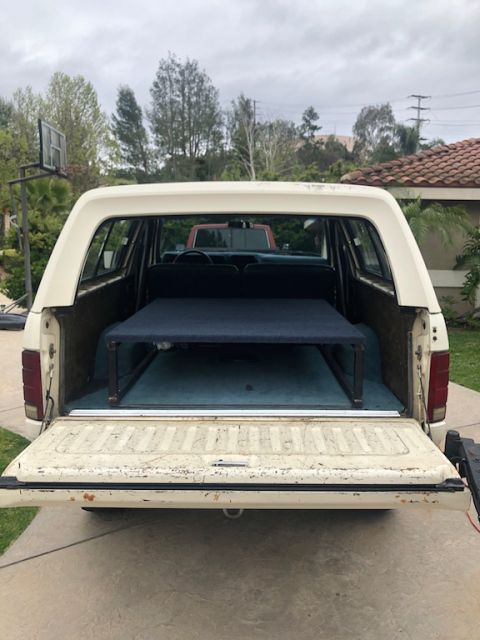 1981 Ford Bronco - Rear view of a cream-colored Ford Bronco with a bed platform, with a red truck visible in the background.