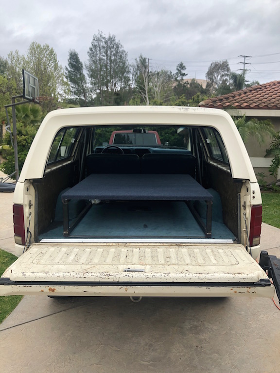 Cream-colored Ford Bronco with tailgate down, showing a raised bed platform inside. A red truck is visible behind it
