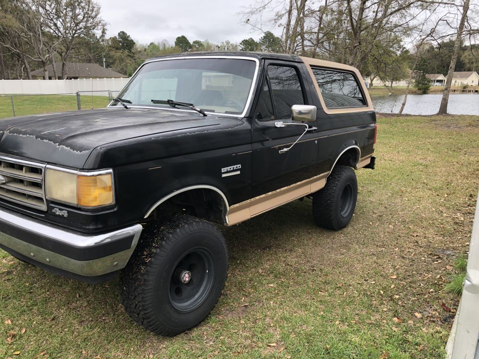Black 1987 Ford Bronco with tan removable top and tan side trim, lifted on black wheels, parked on grass
