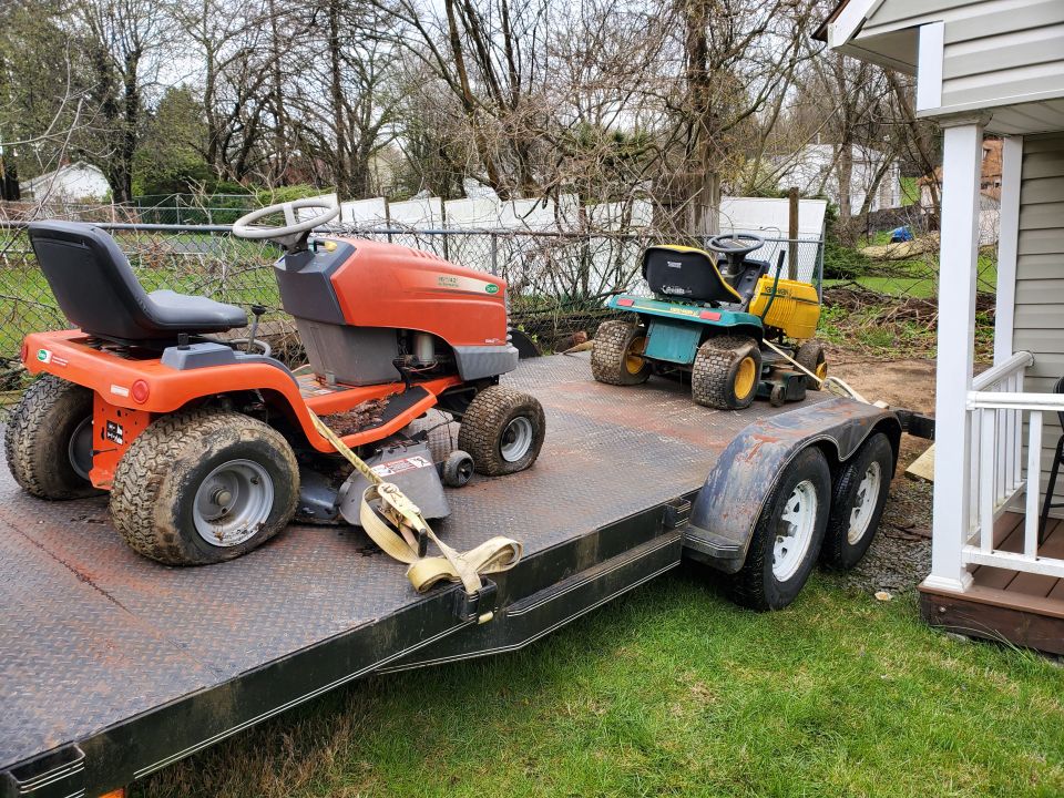 Two riding lawn mowers on a trailer, one orange, one green and yellow