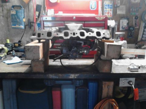 1978 Ford Bronco - An engine intake manifold is disassembled and resting on wooden blocks on a workbench in a garage setting.