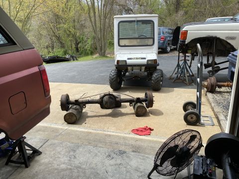 1994 Ford Bronco - A rear axle assembly sits on logs in a garage setting, with a maroon SUV and a white utility vehicle visible.