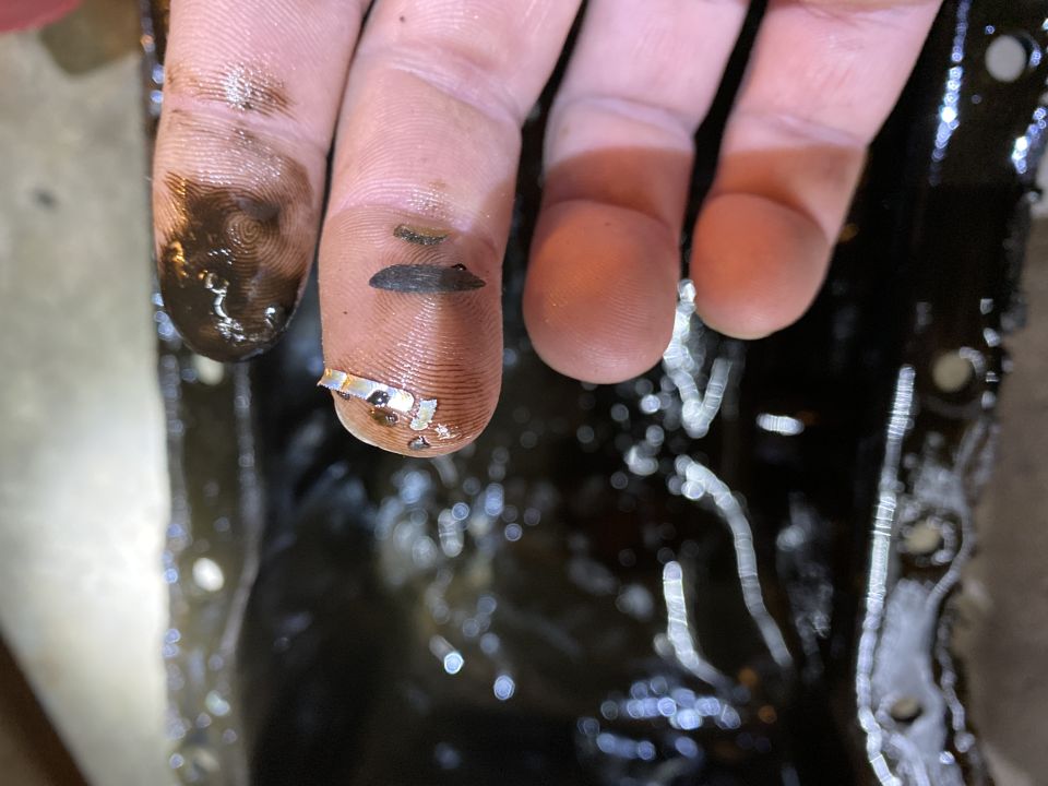 Fingers covered in oil with metal shavings on them, resting above a dark, oily surface that appears to be an oil pan