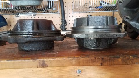 1985 Ford Bronco - Two metal automotive parts, possibly hub assemblies, sit on a wooden workbench. The part on the left is darker and appears older or dirtier…