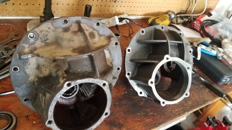 1985 Ford Bronco - Two differential housings are laid out on a wooden workbench. The housing on the left is larger and appears to be older, with some grease a…