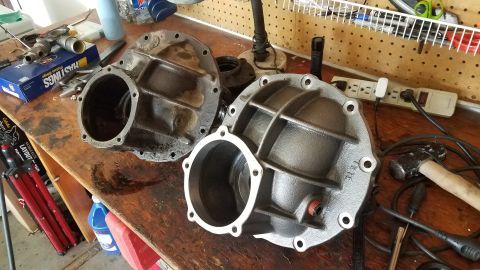 1985 Ford Bronco - Two differential housings are on a wooden workbench. The housing on the right is freshly painted gray and has fins for structural support.…