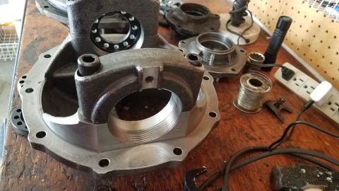 1985 Ford Bronco - A close-up view of a differential housing and related parts laid out on a wooden workbench. Several metal components, including bearings an…