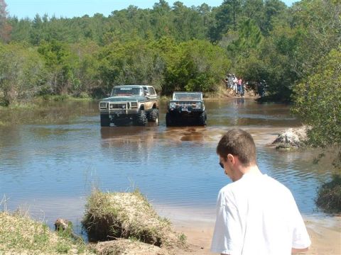 1986 Ford Bronco - Two modified SUVs navigate a water crossing, with onlookers gathered on the bank in the distance.