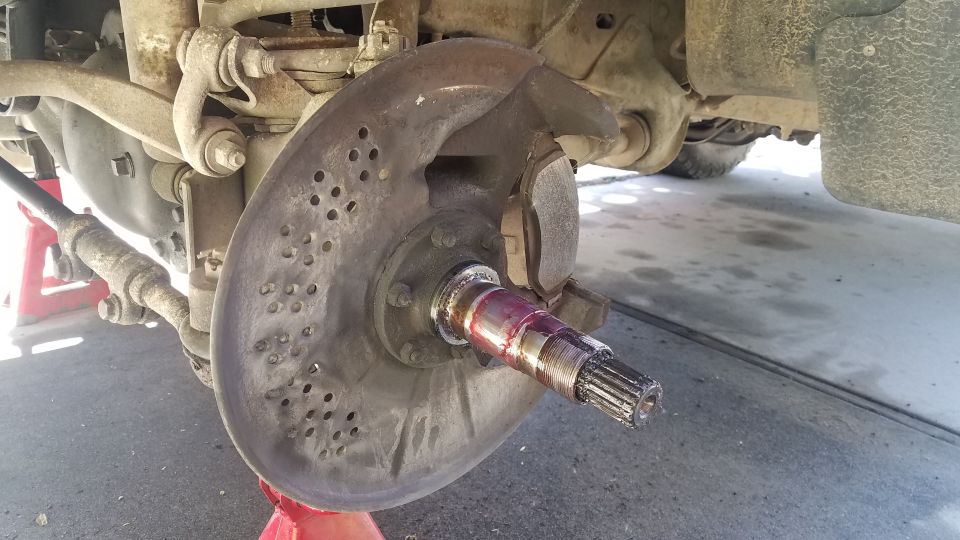 Close-up of a vehicle's front wheel hub with a brake rotor and suspension components visible. The hub is greased and the rotor has many small holes