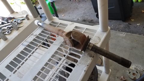 1985 Ford Bronco - A rusty driveshaft with a universal joint is laid across a white plastic shelving unit. Tools are visible on the shelf to the left.