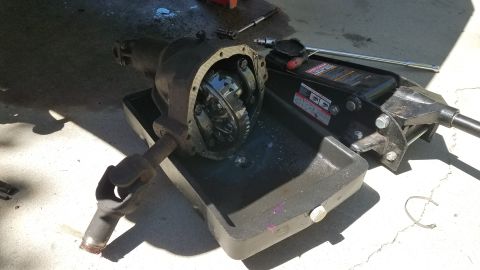 1985 Ford Bronco - A differential housing with the cover removed, showing the internal gears, rests in a black oil drain pan next to a hydraulic floor jack an…