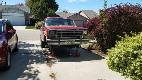 1985 Ford Bronco - A red 1985 Ford Bronco is parked on a driveway, supported by red jack stands. A dark red SUV is parked to the left of the Bronco.
