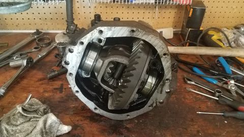 1985 Ford Bronco - A differential housing is open on a wooden workbench, revealing the internal gears. Various tools, including wrenches and pliers, are scatt…
