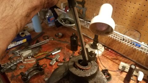 1985 Ford Bronco - A person's hand is holding a tool over a greasy, dark-colored automotive part, possibly a differential housing, on a workbench cluttered wi…
