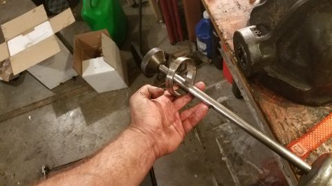 1985 Ford Bronco - A person's greasy hand holds a metal tool with a knurled knob and a ring, preparing to work on a dark metal object resting on a wooden work…