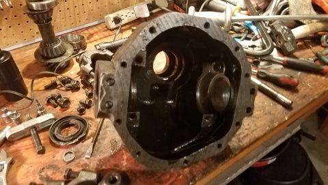 1985 Ford Bronco - A differential housing is disassembled on a workbench with various tools and parts scattered around it. The housing is dark and appears to…