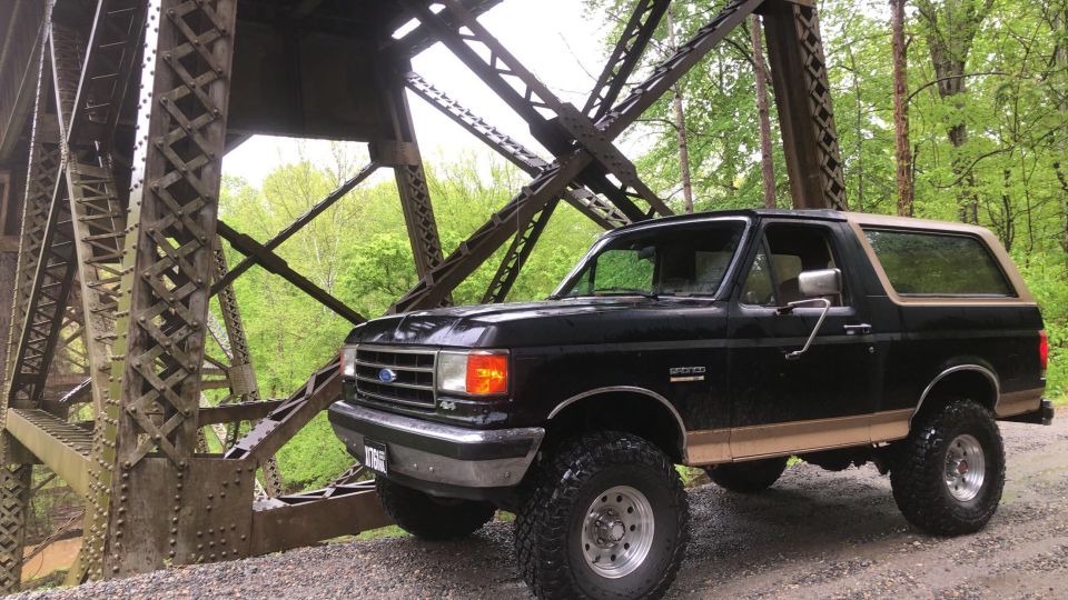 Black 1991 Ford Bronco with tan stripe, lifted, off-road tires, parked under a metal bridge.
