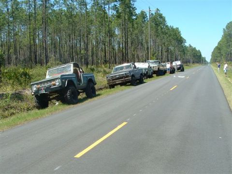 1986 Ford Bronco - A line of off-road vehicles parked along a rural road next to a dense forest.